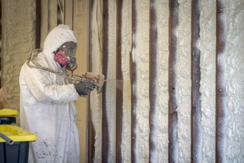 Person standing and spraying spray foam installation into a wall while wearing PPE